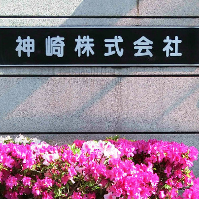 グレーの壁に黒い背景で「神崎株式会社」と記した銘板。その下には赤やピンクのつつじがたくさん咲いています。
