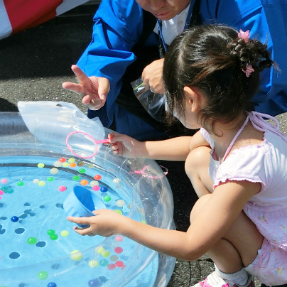 透明のビニルプールにスーパーボールをたくさん浮かべています。小さな女のお子さんがポイをもって救っています。