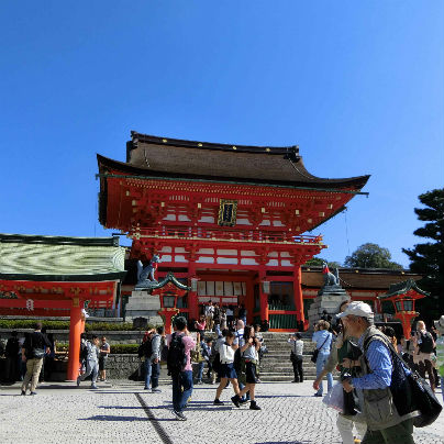 真っ青な空を背景に伏見稲荷大社の朱い楼門、数十人の参拝客が映っています。