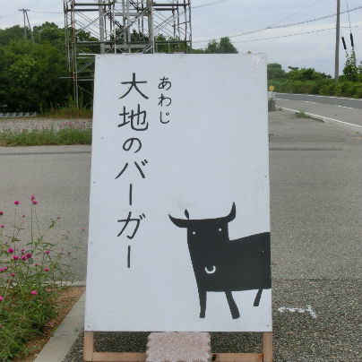 白い立て看板が道に一つ。黒い文字で「あわじ大地のバーガー」と書いてあり、右下には正面を向いた牛のイラストがあります、