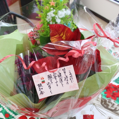 クリスマスカラーのテーブルクロスの上に赤いポインセチアをメインにしたアレンジメント。メッセージカードにには「この花に皆様のご健康とお幸せの願を込めて  阪和会」と書かれています。