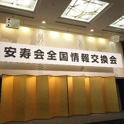 金屏風を背景に天井から白い横長の板に黒い文字で安寿会全国情報交換会と書いてあります。