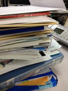 白い机の上にファイルやらカタログやら封筒やらが積み上がっています