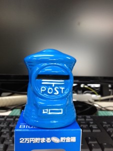 白い机の上に、パソコンモニターを背景に青色の郵便ポスト型の貯金箱が置いてあります。
